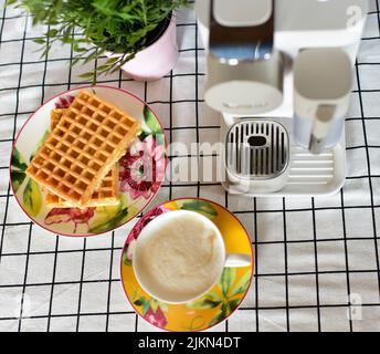 Ein Blick von oben auf eine Tasse Kaffee, Waffeln, Kaffeemaschine und Pflanzen auf einer karierten Tischdecke Stockfoto