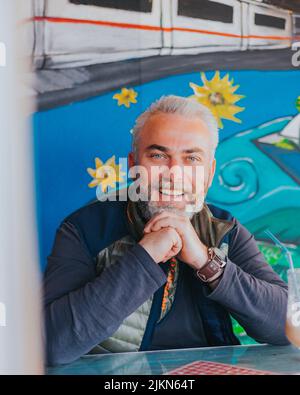Eine vertikale Aufnahme eines hübschen Erwachsenen mit grauen Haaren und blauen Augen, der glücklich in einem Café sitzt Stockfoto