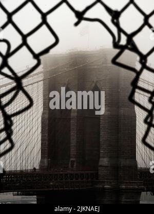 Eine vertikale Graustufenaufnahme der Brooklyn Bridge, aufgenommen aus einem Loch in einem Kettenzaun Stockfoto