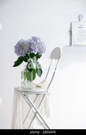 Die vertikale Studioaufnahme der Stillleben Komposition mit einem weißen Stuhl und einer Vase mit violetten Blumen Stockfoto
