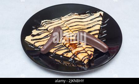 Eine Nahaufnahme von Crepes mit Schokoladensirup und Süßigkeiten auf einem schwarzen Teller Stockfoto