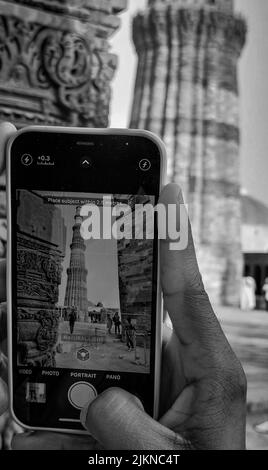 Eine Graustufe der Hand einer Person mit einem Telefon, das ein Foto eines alten Qutab Minar Turms macht Stockfoto