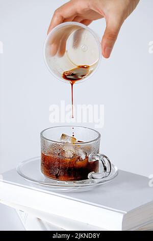 Eine vertikale Aufnahme einer Hand, die einer Tasse mit Eiswürfeln auf weißem Hintergrund allmählich Kaffee hinzufügt Stockfoto