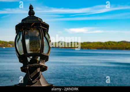 Eine Nahaufnahme einer alten Straßenlaterne am See und grünen Laubbäumen am Horizont gegen den blauen Himmel Stockfoto