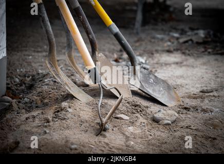 Schmutzige gebrauchte Schaufeln und ein Rechen lehnen sich während einer Arbeitspause auf einer neuen Baustelle für den Hausbau an eine Wand. Stockfoto