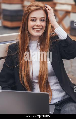 Lächelnde, rothaarige, schöne Geschäftsfrau, die mit einem Laptop auf einer Holzbank im Park arbeitet. Positive Person. Kinder Lifestyle Kindheitskonzept. Stockfoto