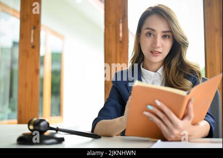 Charmante junge asiatische Rechtsanwältin oder Business-Rechtsberaterin mit ihrem Anwaltsbuch und sitzt an ihrem Schreibtisch. Stockfoto