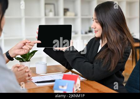 Eine attraktive junge asiatische Immobilienmaklerin, die einen digitalen Tablet-Bildschirm hält, einen schwarzen Bildschirm zeigt und etwas auf dem Tablet-Bildschirm präsentiert. Stockfoto