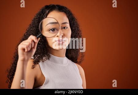 Lassen Sie mich einen genaueren Blick darauf werfen. Ein beschnittenes Porträt einer attraktiven jungen Frau, die im Studio vor einem roten Hintergrund durch eine Lupe schaut. Stockfoto