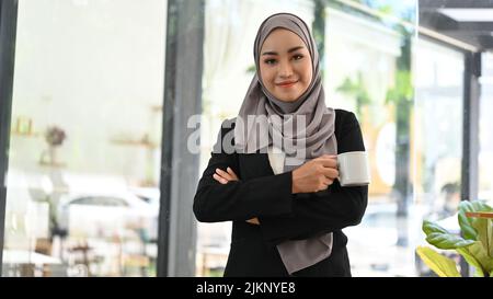 Schöne junge asiatische muslimische Geschäftsfrau oder weibliche Chefin in einem formellen Anzug und Hijab steht vor ihrem Büro mit einer Tasse Kaffee in ihren Händen. Stockfoto