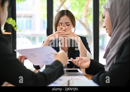 Gestresste und müde, tausendjährige asiatische Chefin ist bei einem Treffen mit ihrem Team, unzufrieden mit ihrem Geschäftsumsatz, während sie zuhört Stockfoto