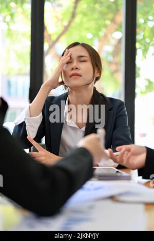 Gelangweilte und müde junge asiatische Geschäftsfrau oder Managerin bekam während des Finanzmeeting mit ihrem Team Kopfschmerzen. Frustrierte Geschäftsfrau Stockfoto