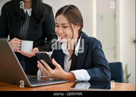 Fröhliche und fröhliche junge asiatische Geschäftsfrau oder Büroangestellte an ihrem Schreibtisch im Gespräch mit ihrem Kollegen. Stockfoto