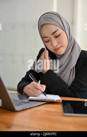 Eine hochporträtierte, attraktive und professionelle junge asiatische muslimische Geschäftsfrau oder Sekretärin mit Hijab sitzt an ihrem Schreibtisch und macht sich Notizen Stockfoto