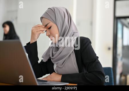 Gestresste, tausendjährige asiatische muslimische Geschäftsfrau oder Büroangestellte, die an ihrem Schreibtisch arbeitete, frustriert über ihr Projekt, an einem Kopf leidend Stockfoto