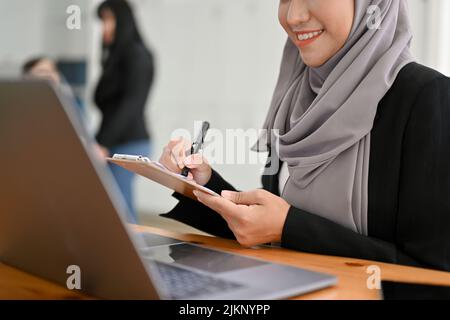 Eine schöne junge asiatische muslimische Geschäftsfrau oder Büroangestellte, die an ihrem Schreibtisch arbeitet, auf den Laptop-Bildschirm schaut und etwas auf ihrem Notizblock notiert Stockfoto