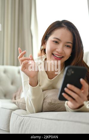 Porträt, attraktive und glückliche junge asiatische Frau, die auf ihrem bequemen Sofa liegt, in den sozialen Medien scrollt und mit ihren Freunden auf ihrem Smartphone chattet. Stockfoto