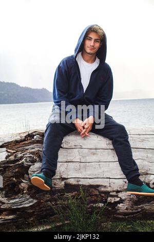Eine helle vertikale Aufnahme eines kaukasischen Mannes, der einen Hoodie trägt und auf einem Holzbalken vor dem See sitzt Stockfoto