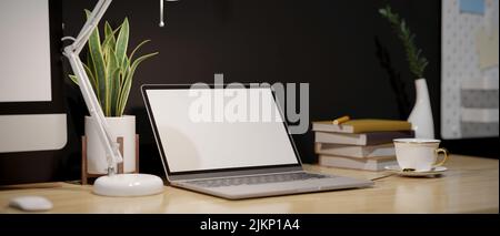 Nahaufnahme, moderner Arbeitsplatz mit tragbarem Laptop-Modell mit weißem Bildschirm, Tischlampe, Kaffeetasse, Buch und Dekors auf Holztisch gegen den schwarzen Wal Stockfoto