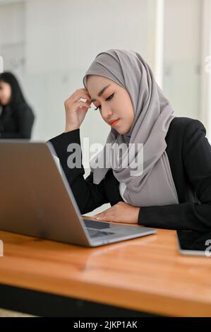 Gestresste junge asiatische muslimische Geschäftsfrau oder Geschäftsangestellte, die an ihrem Schreibtisch vor dem Laptop arbeitet. Stockfoto