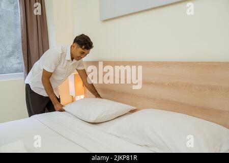 Junge männliche Haushälterin, die im Hotelzimmer Kissen auf dem Bett aufräumte Stockfoto