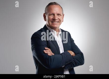 Ein Spielwechsel in einer Welt voller Spieler. Ein gutaussehender, reifer Geschäftsmann, der allein vor grauem Hintergrund im Studio mit gefalteten Armen steht. Stockfoto