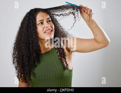 Das Kämmen meiner Haare ist nie eine leichte Aufgabe. Eine Frau runzelte die Stirn, während sie ihr Haar vor einem grauen Hintergrund kämmte. Stockfoto