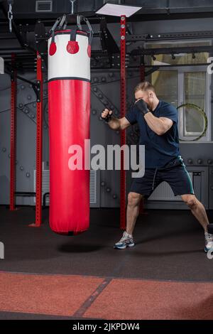 Junger starker bärtiger Mann, Kämpfer und Trainer der gemischten Kampfkunst, trainiert im Fitnessstudio. Steht in defensiver Haltung vor einem roten Boxsack. Stockfoto