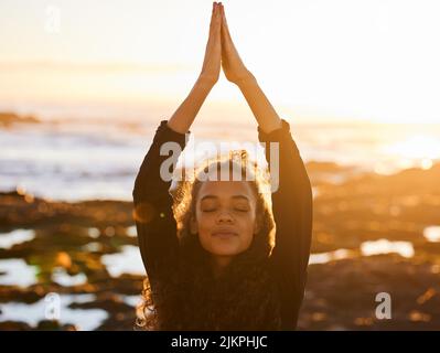 Sammeln Sie gute Energie aus dem Universum. Eine attraktive junge Frau, die beim Sonnenuntergang am Strand Yoga macht. Stockfoto