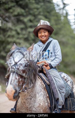 27.07.2022, Kirgisistan, Issyk-Kul. Ein süßer, lächelnder asiatischer Junge in einem Nationalhut reitet auf einem Pferd in den Bergen und trifft Touristen. Nahaufnahme. Stockfoto