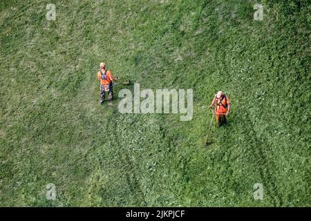 Zwei Arbeiter mit einem Rasenmäher mähen das Gras, Draufsicht. Mans in einer orangefarbenen Arbeitsuniform schneidet Gras Stockfoto