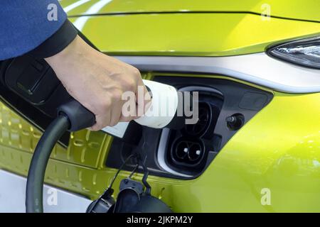 Nahaufnahme der Hand eines Mannes, der einen wiederaufladbaren Gerätestecker für die Ladebatterie an der Buchse eines weißen Elektroautos hält Stockfoto