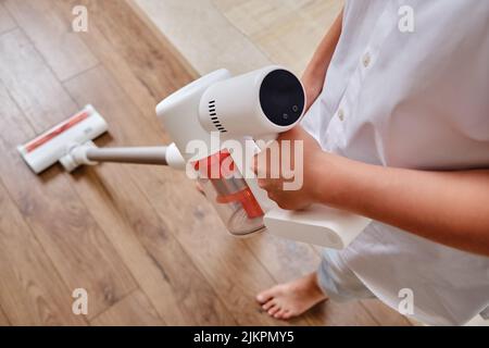 Hände und Display von Frauen steuern den kabellosen tragbaren Staubsauger in der Küche. Eine Frau reinigt den Boden in einer Wohnung mit einem Staubsauger mit einem Stockfoto