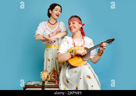 Musikerinnen in russischen Volkskleidern mit Musikinstrumenten auf blauem Studiohintergrund. Glückliche Künstler aus Russland in weißer Nationalkleidung mit Stockfoto