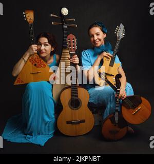 Musikerinnen in blauen Kleidern mit Musikinstrumenten auf schwarzem Studiohintergrund. Fröhliche Künstler mit Saiteninstrumenten auf dunklem Backg Stockfoto