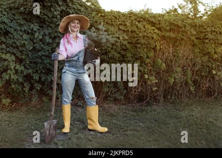 Eine junge Frau pflanzt einen Weihnachtsbaum in den Boden. Das Konzept der Landschaftsgestaltung, Gartenarbeit - ein Gärtner pflanzt Bäume in einem Sommergarten. Stockfoto