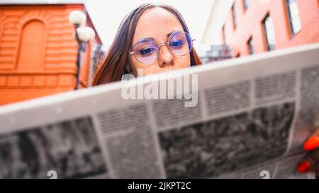 Nachdenkliche junge Frau, die Zeitung auf der Straße liest. Verwirrt junge Unternehmerin mit langen braunen Haaren in formeller Kleidung und Brillen stehend Stockfoto
