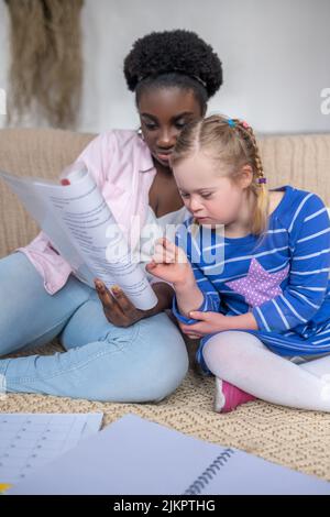 Dunkelhäutige Frau, die einem Mädchen mit Down-Syndrom etwas vorliest Stockfoto