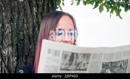 Konzentrierte elegante Dame beim Lesen der Zeitung im Park. Fokussierte junge Unternehmerin mit langen, farbstoffgefärbten Haaren in formeller Kleidung und Brillen, die in der Nähe stehen Stockfoto