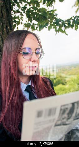 Konzentrierte elegante Dame beim Lesen der Zeitung im Park. Fokussierte junge Unternehmerin mit langen, farbstoffgefärbten Haaren in formeller Kleidung und Brillen, die in der Nähe stehen Stockfoto