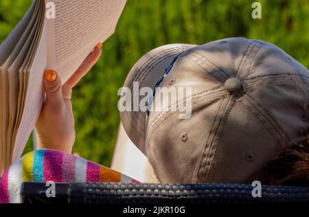 Nahaufnahme einer jungen Dame mit orangefarbenen Fingernägeln, die eine Baseballmütze trägt und ein Taschenbuch liest, während sie im Urlaub auf einer Sonnenliege faulenzt Stockfoto