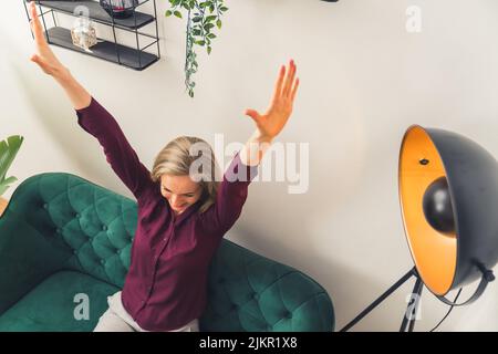 Kaukasische Frau mit blonden Haaren trägt kastanienbraune Hemd entspannen auf grünem Sofa halten die Arme nach oben Stretching. Innenaufnahme. . Hochwertige Fotos Stockfoto