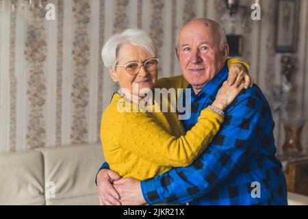 Liebevolles altes, verliebtes Ehepaar aus der älteren Familie lächelt und umarmt sich im Wohnzimmer zu Hause und genießt ihren Ruhestand. Hochwertige Fotos Stockfoto