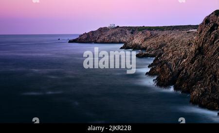 Felsige Küste von Menorca, Spanien, in der Abenddämmerung, mit einem kleinen weißen Haus auf der Klippe, ruhigem Mittelmeer und sanftem rosa Abendhimmel auf den Balearen ist Stockfoto
