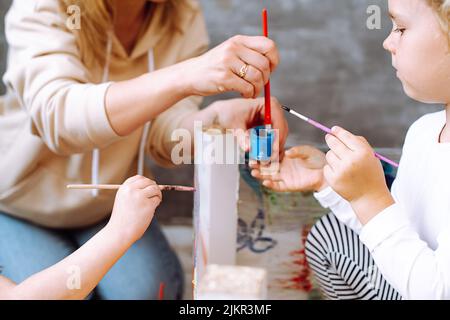 Zugeschnittenes Foto von jungen Lehrerin, die zeigt, wie man blaue Gouache mit rotem Pinsel im Plastikglas zu kleinen Mädchen rühren kann. Stockfoto