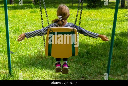 Das kleine Mädchen sitzt mit dem Rücken auf einer Schaukel und reitet im Sommer Stockfoto