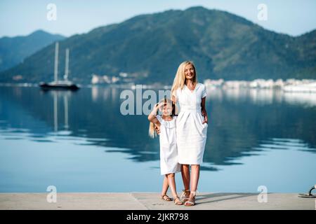 Glückliche, tausendjährige Mutter und blonde Teenager-Tochter in weißen Kleidern im Familienlook, die am Hafen und an der Küste in einem touristischen Luxusresort am Meer spazieren gehen Stockfoto