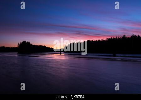 Der Fluss Pielisjoki fließt in der Dämmerung durch die Winterwaldlandschaft. Farbenprächtiger Dämmerungshimmel und seine Spiegelungen im Wasser. Finnland. Stockfoto