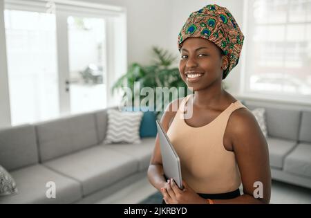 Porträt einer schönen lächelnden afrikanischen Frau, die in einem modernen Zuhause mit einem Laptop in der Hand steht und ein traditionelles Kopftuch trägt Stockfoto