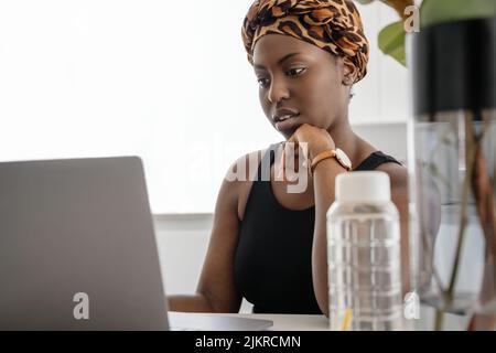 Schöne junge afrikanische Frau trägt Tradition Kopftuch. Sitzen zu Hause und arbeiten auf einem Laptop. Stockfoto